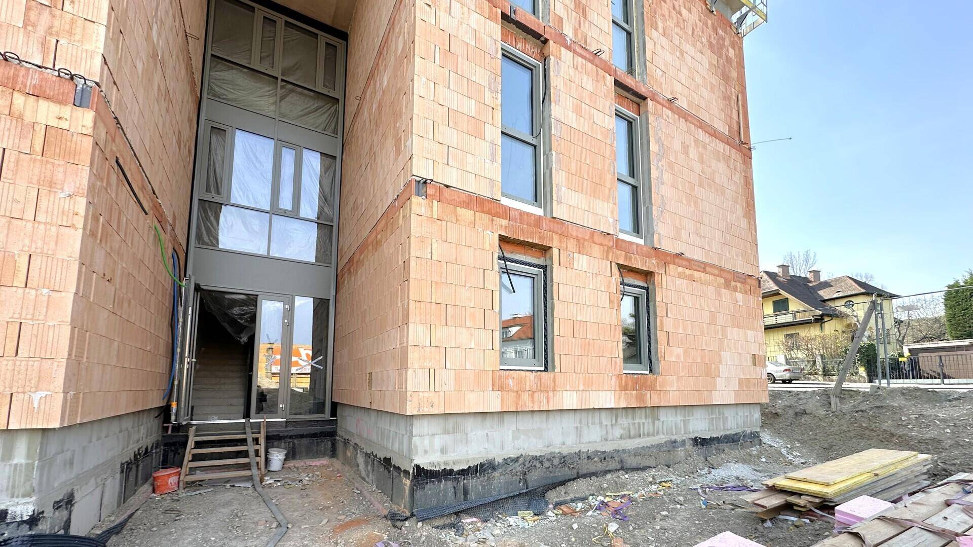 Eingangsbereich eines Neubaus im Rohbau mit großen Fenstern und provisorischer Treppe.