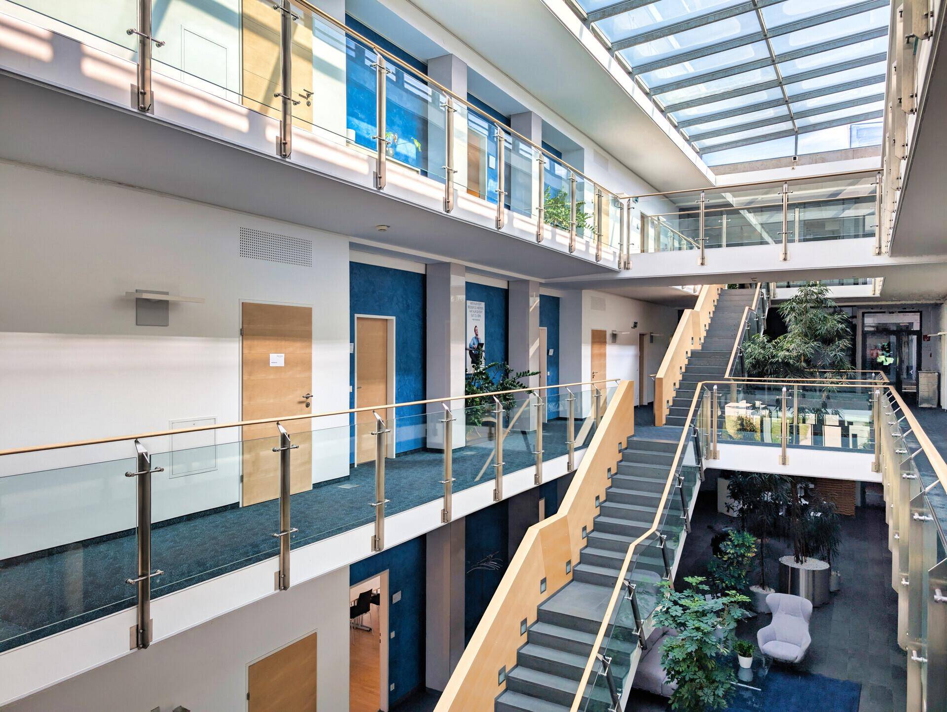 Mehrstöckiges Atrium eines Bürogebäudes mit Glasdach, Treppen, Glasgeländern und Pflanzen, das viel Tageslicht bietet.