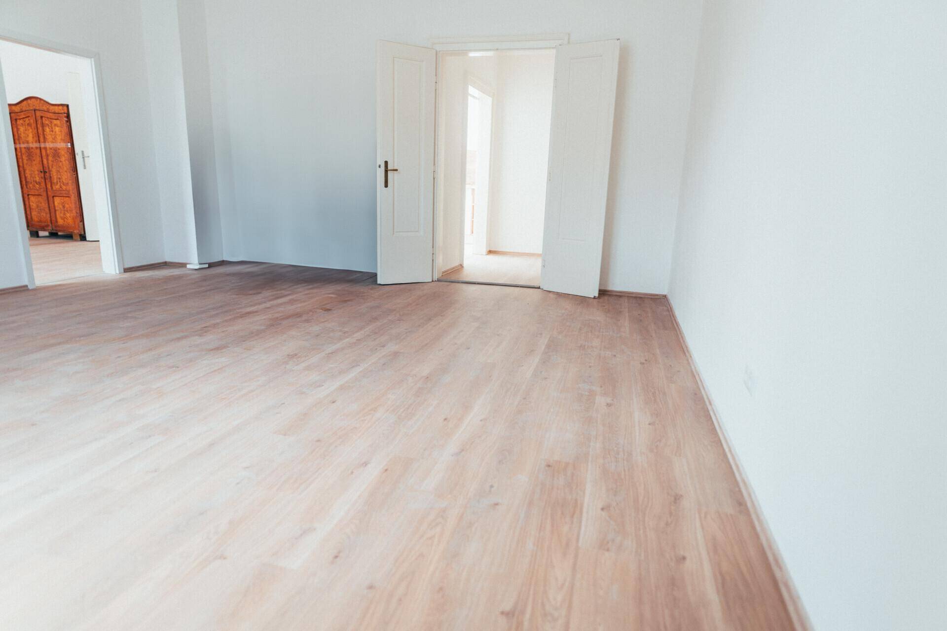 Heller Raum mit Laminatboden und weißen Wänden, offene Türen führen zu einem angrenzenden Zimmer.
