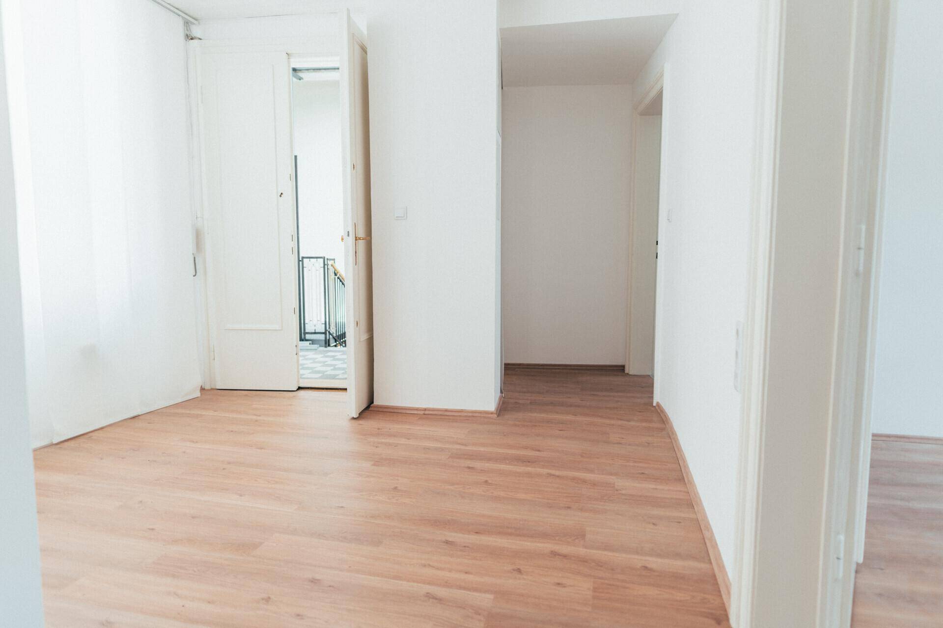 Heller, leerer Raum mit Laminatboden und weißen Wänden, offene Türen führen zu einem angrenzenden Bereich.