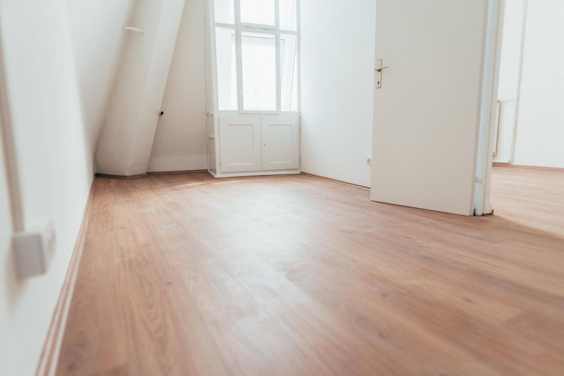 Leerer Raum mit Dachschräge, hellem Laminatboden und einem Fenster, das viel Tageslicht hereinlässt.