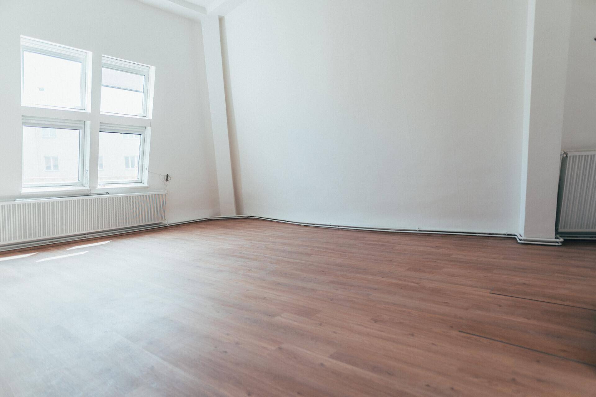 Geräumiger, heller Raum mit Laminatboden, weißen Wänden und einem Fenster mit Heizkörper.