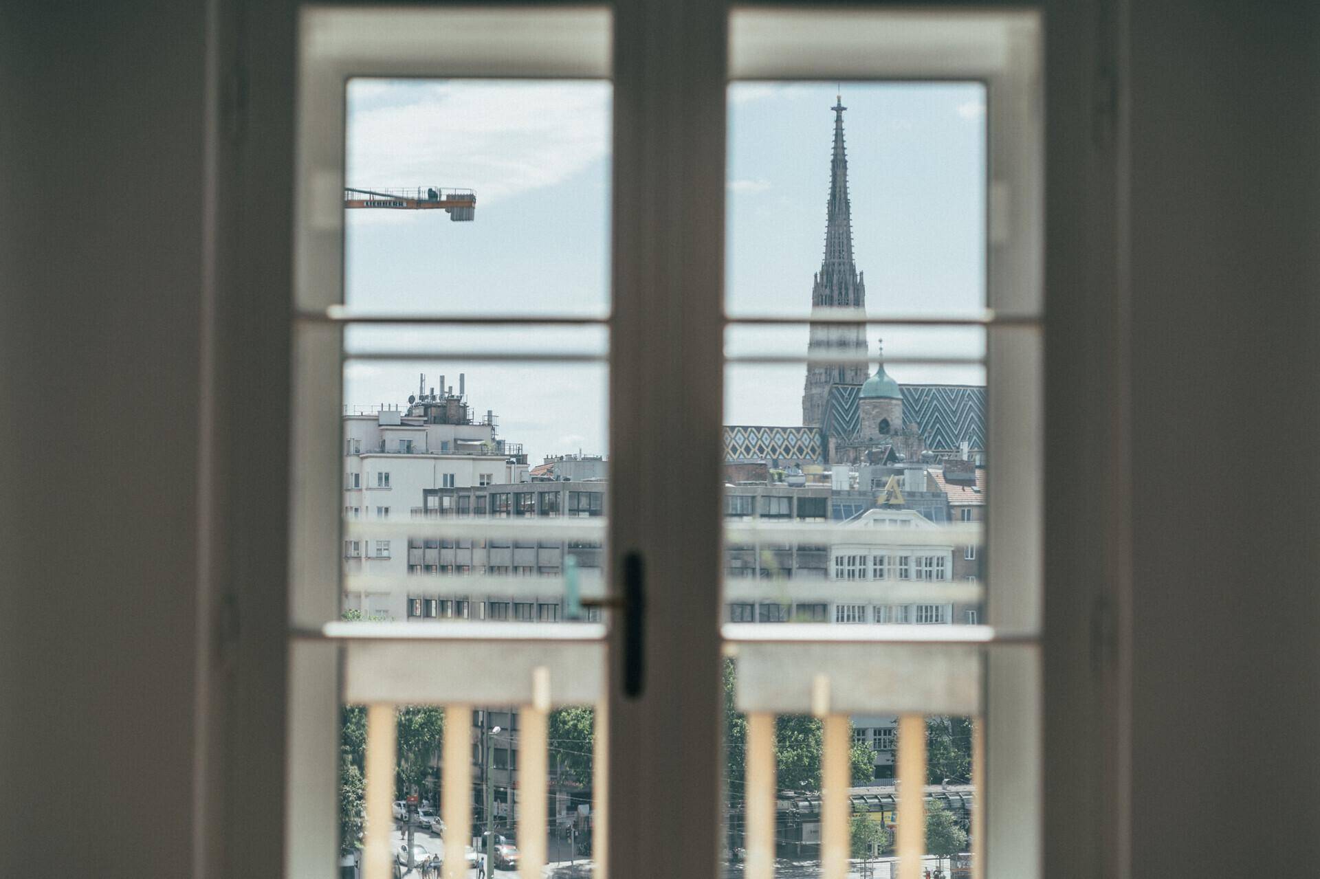 Blick aus dem Fenster auf die Stadt mit einer Kirchturmspitze und einem Baukran am Horizont.