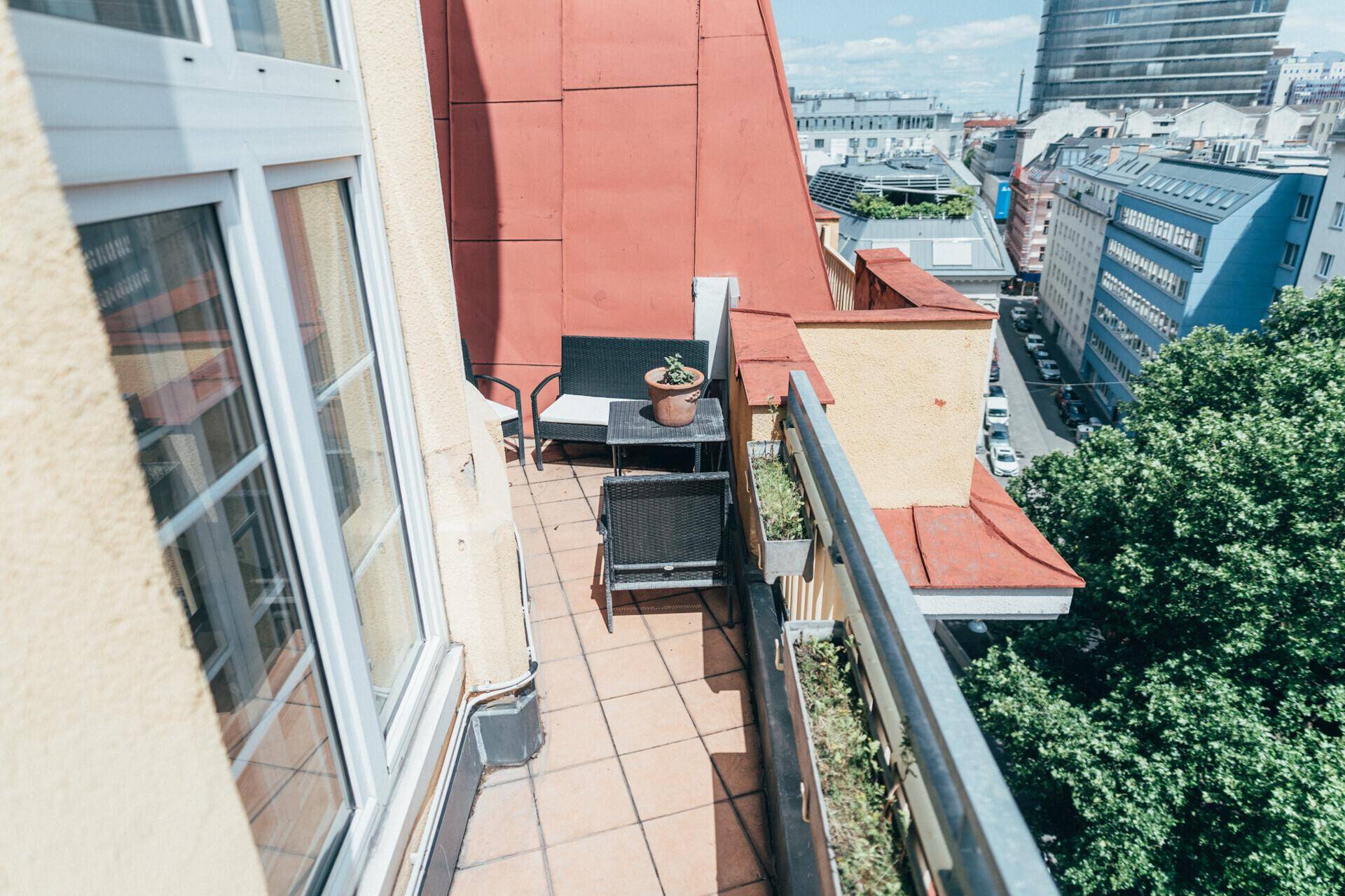 Sonniger Balkon mit Fliesenboden und Sitzgelegenheiten, bietet einen Blick über die Stadt und Bäume.