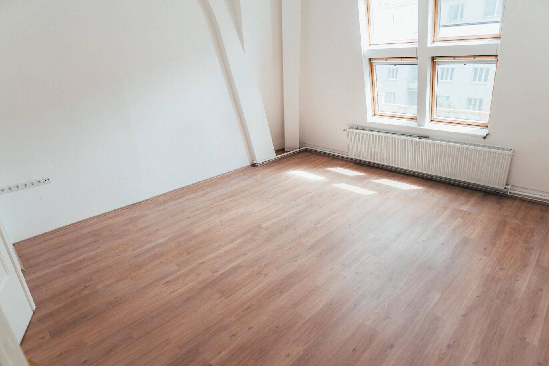 Heller, leerer Raum mit Laminatboden, weißen Wänden und einem Fenster mit Heizkörper darunter.