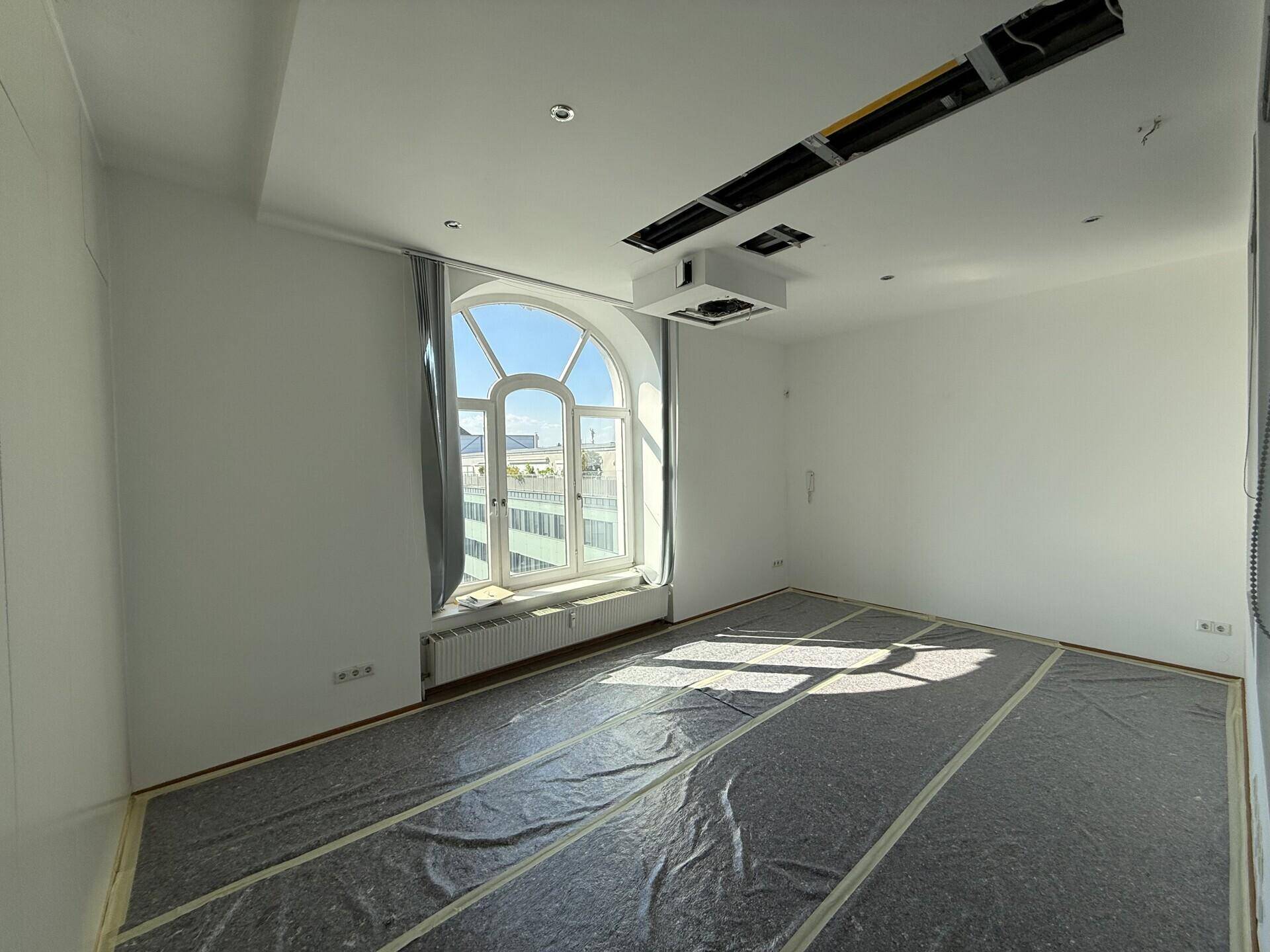 Heller, leerer Raum mit einem großen Rundbogenfenster, Heizkörper und Bodenschutzfolie während des Umbaus.