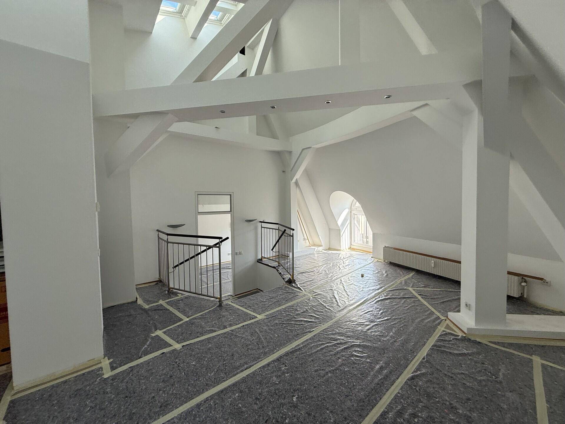 Weitläufiger Dachgeschossraum mit markanten Holzbalken, schrägen Decken und einer Treppe mit Metallgeländer.