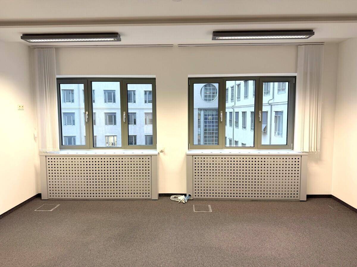 Heller Büroraum mit zwei großen Fenstern, Heizkörperverkleidungen und grauem Teppichboden.
