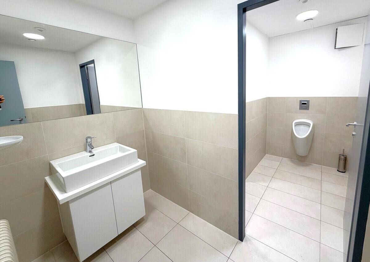 Modernes WC mit Waschbecken, großem Spiegel, Urinal und hellen Fliesen.