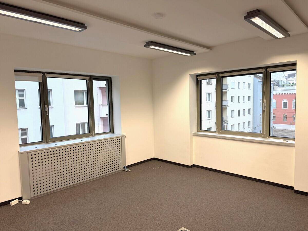 Leerer Büroraum mit großen Fenstern, Heizkörperverkleidungen und grauem Teppichboden.