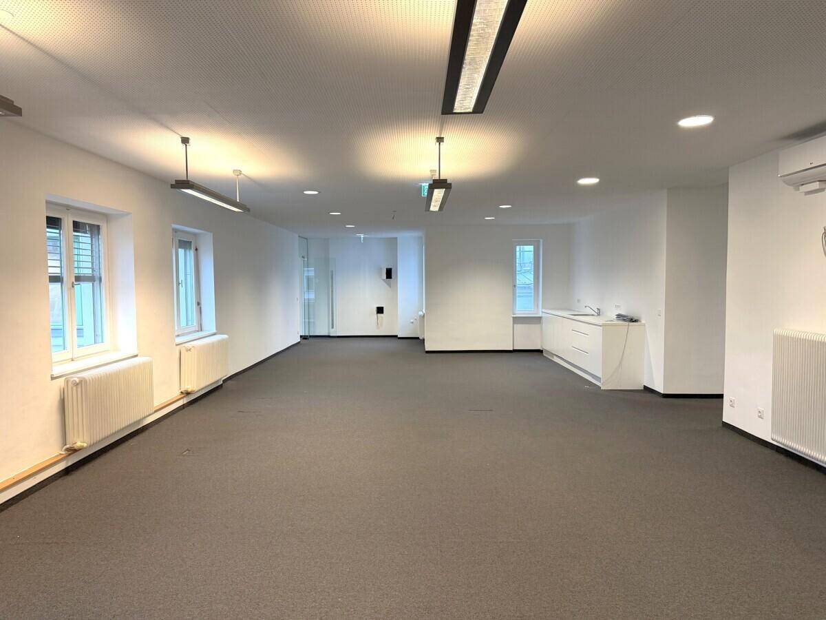 Offener Bürobereich mit Teppichboden, integrierter Küchenzeile und modernen Deckenleuchten.