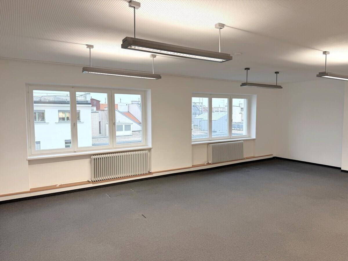 Heller Büroraum mit großen Fenstern, die viel Tageslicht hereinlassen, und einem Blick auf umliegende Dächer.