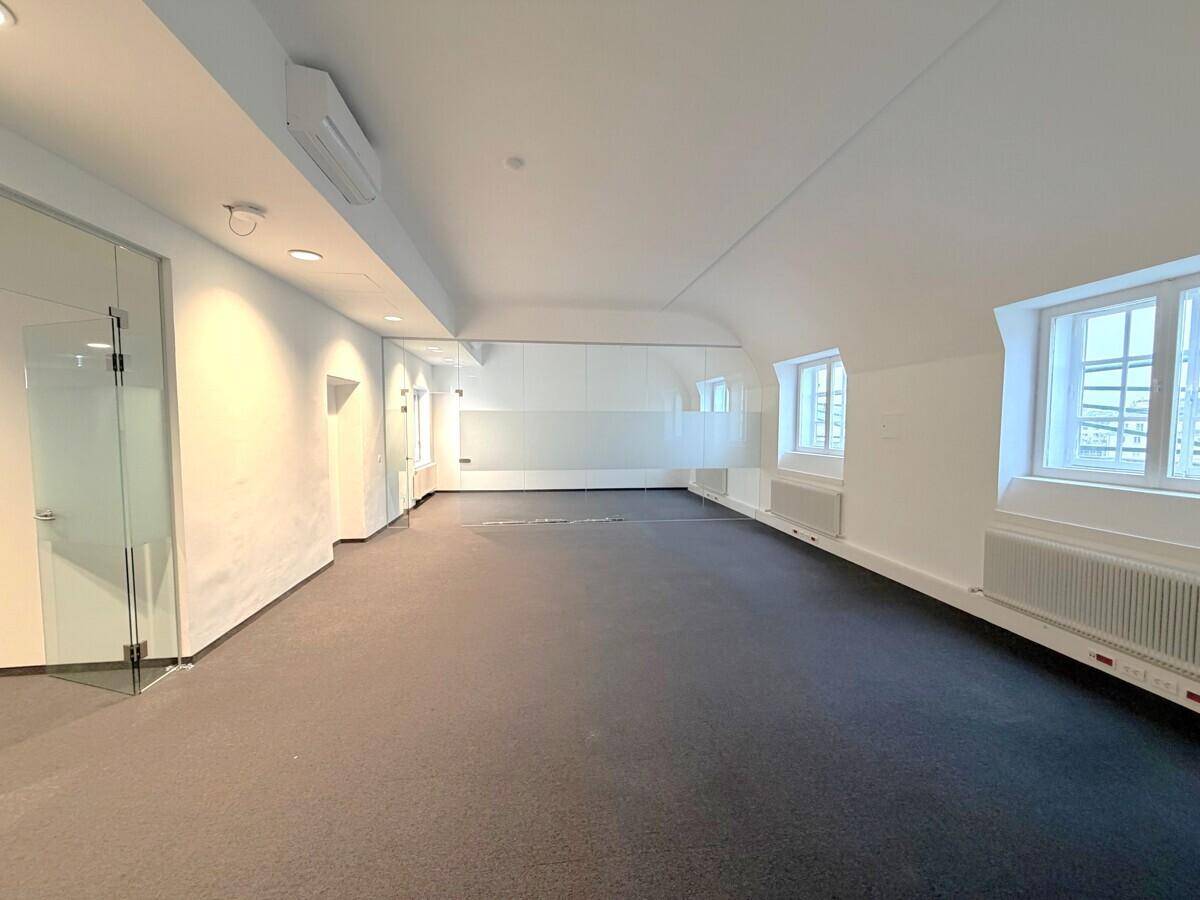 Geräumiger Büroraum mit großen Fenstern, Teppichboden und einer eleganten Glasabtrennung.