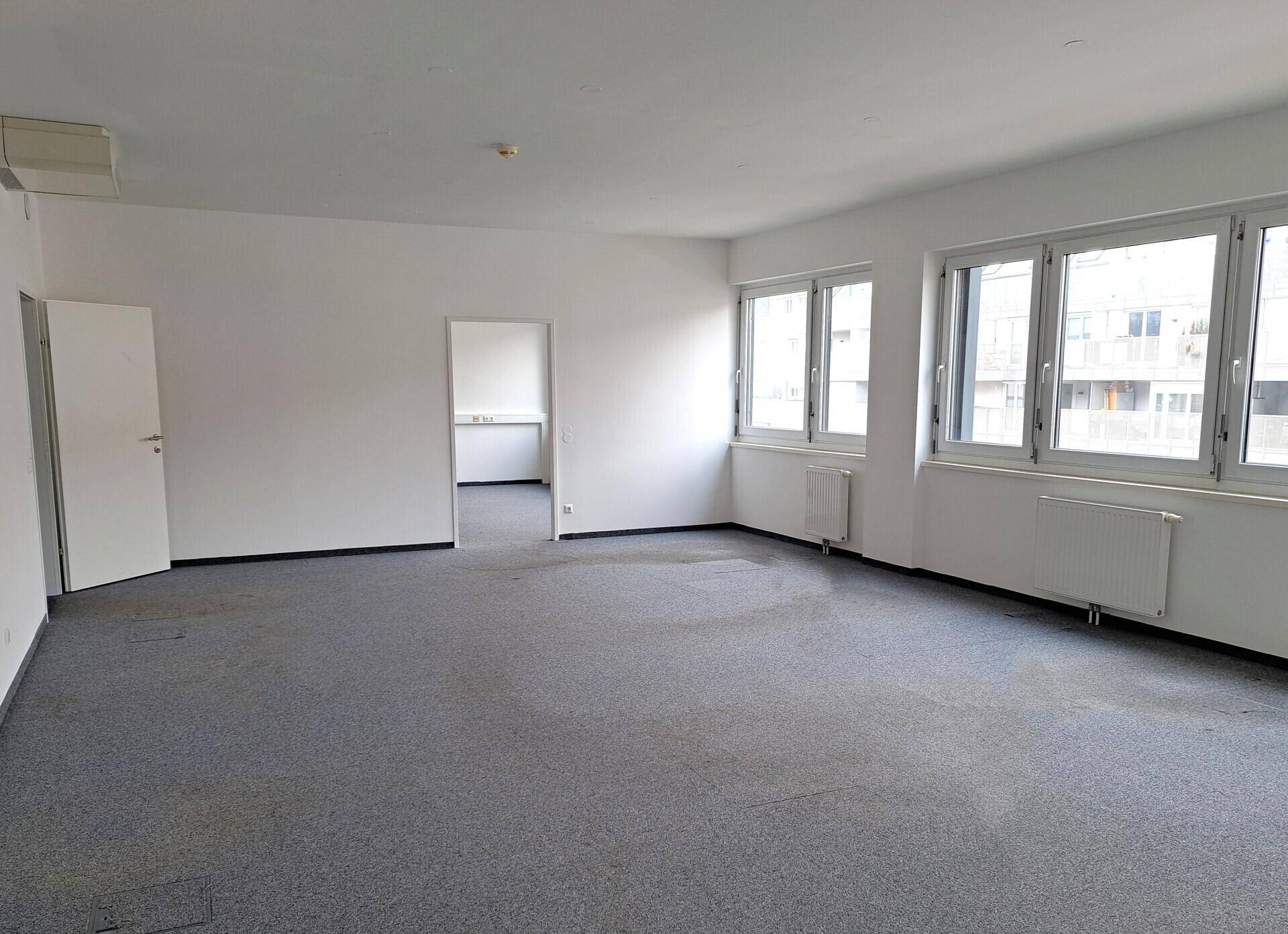 Geräumiger, leerer Büroraum mit grauem Teppichboden, weißen Wänden und großen Fenstern, die viel Tageslicht hereinlassen.