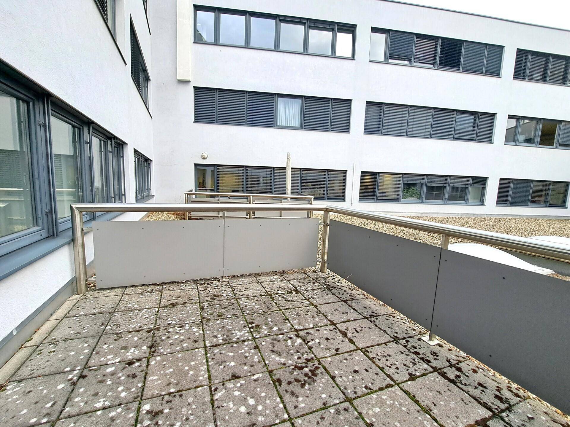 Außenterrasse mit grauen Fliesen und Metallgeländer, umgeben von einer modernen Gebäudefassade mit Jalousien.