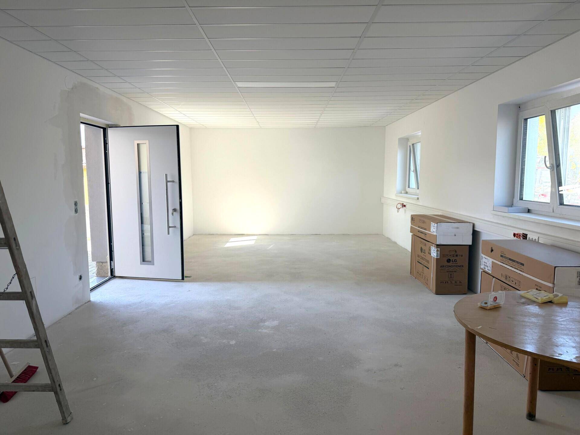 Heller, leerer Raum mit abgehängter Decke, großen Fenstern und einer modernen Eingangstür.