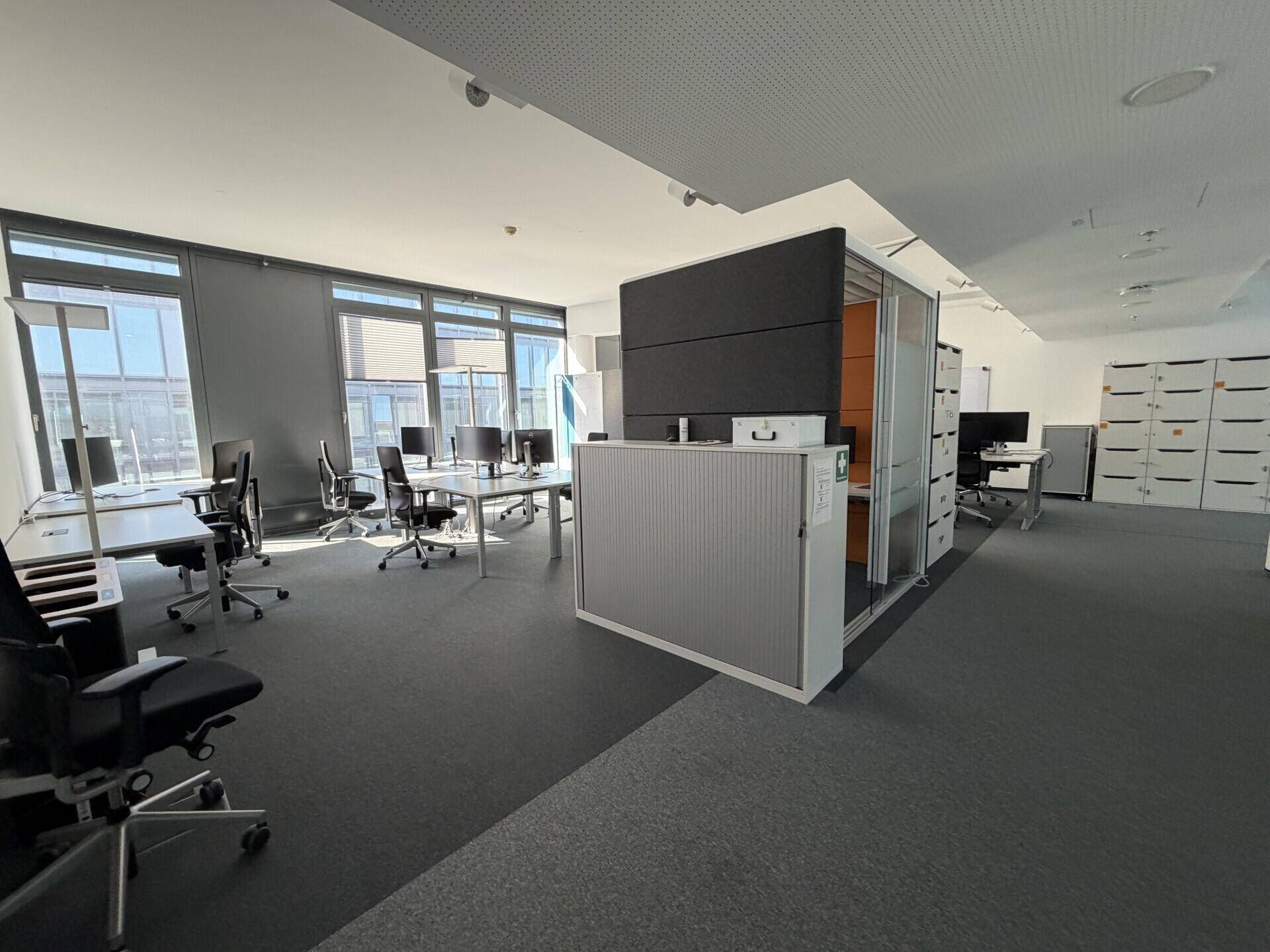 Weitläufiger Bürobereich mit verschiedenen Arbeitszonen, Schließfächern und großen Fenstern.