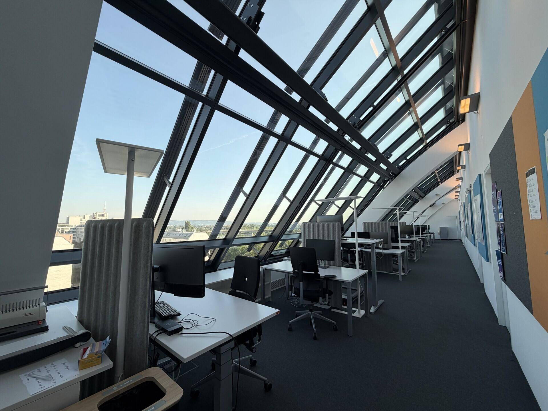 Moderner Büroraum mit vielen Arbeitsplätzen und schrägen Glasfenstern, die viel Tageslicht hereinlassen.