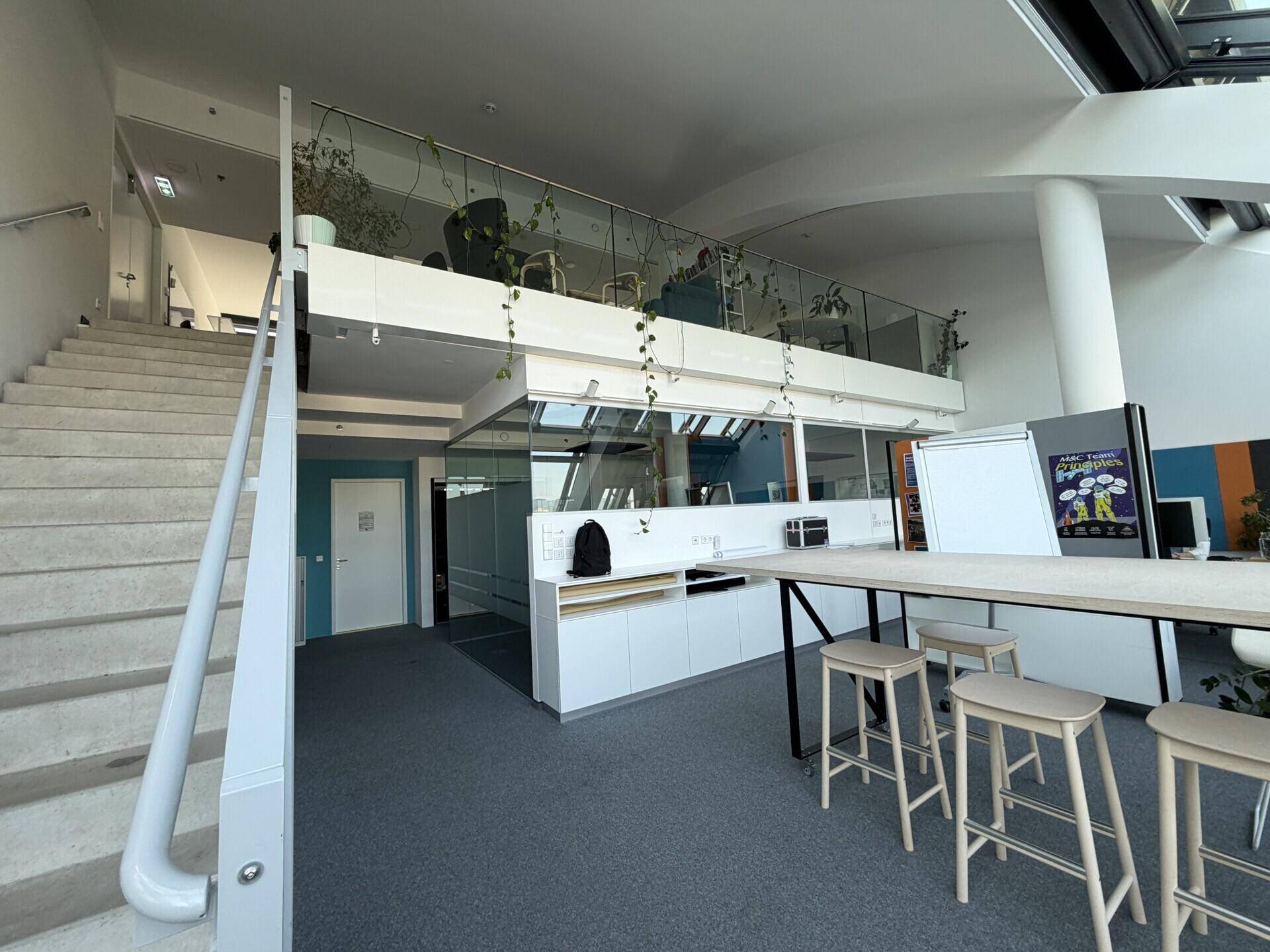Offener Bürobereich mit Treppe zu einer oberen Ebene und einer modernen Küchenzeile im Hintergrund.