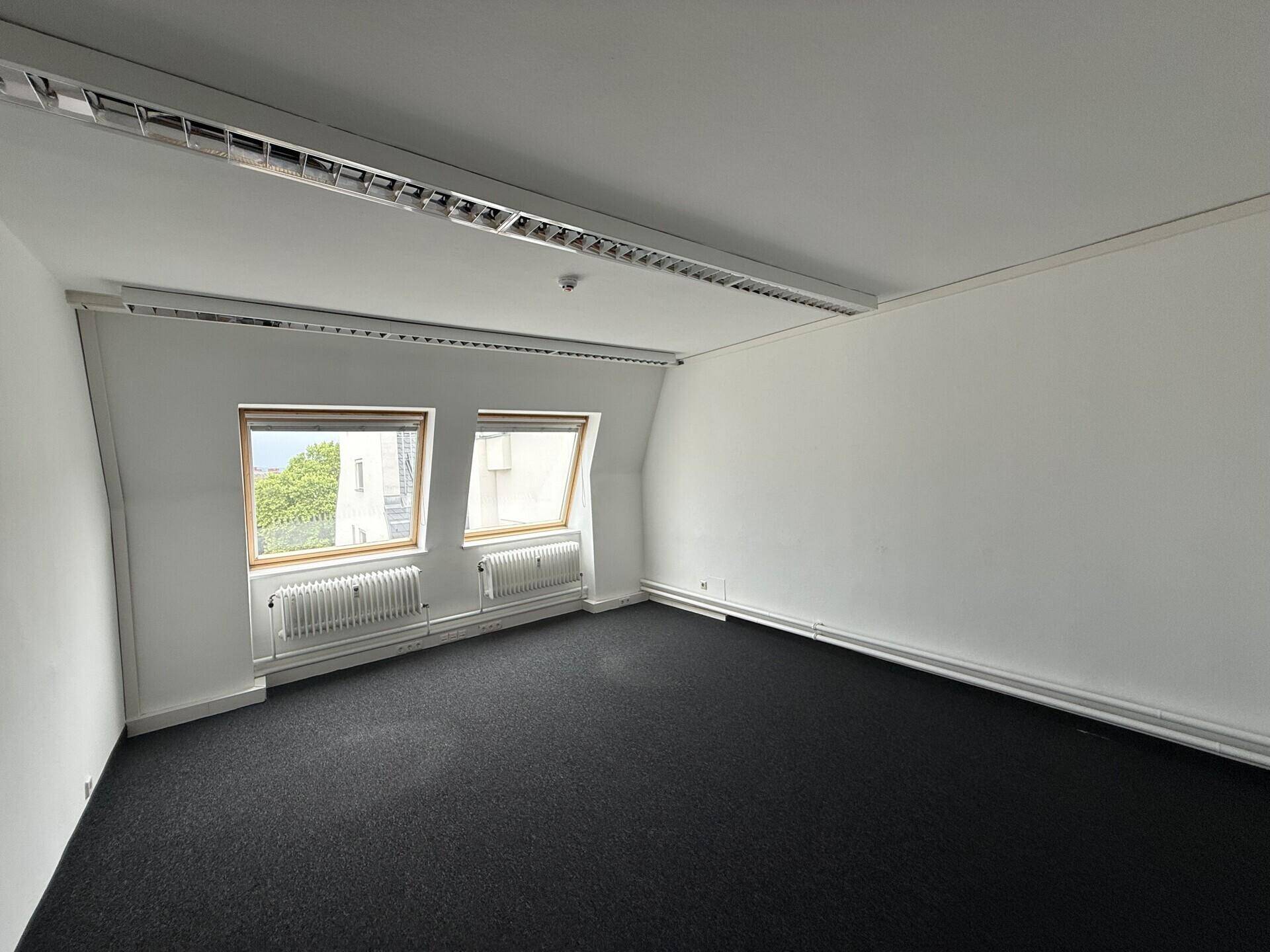 Leerer Raum mit dunklem Teppichboden, schrägen Wänden und zwei Dachfenstern, ideal als Büro.