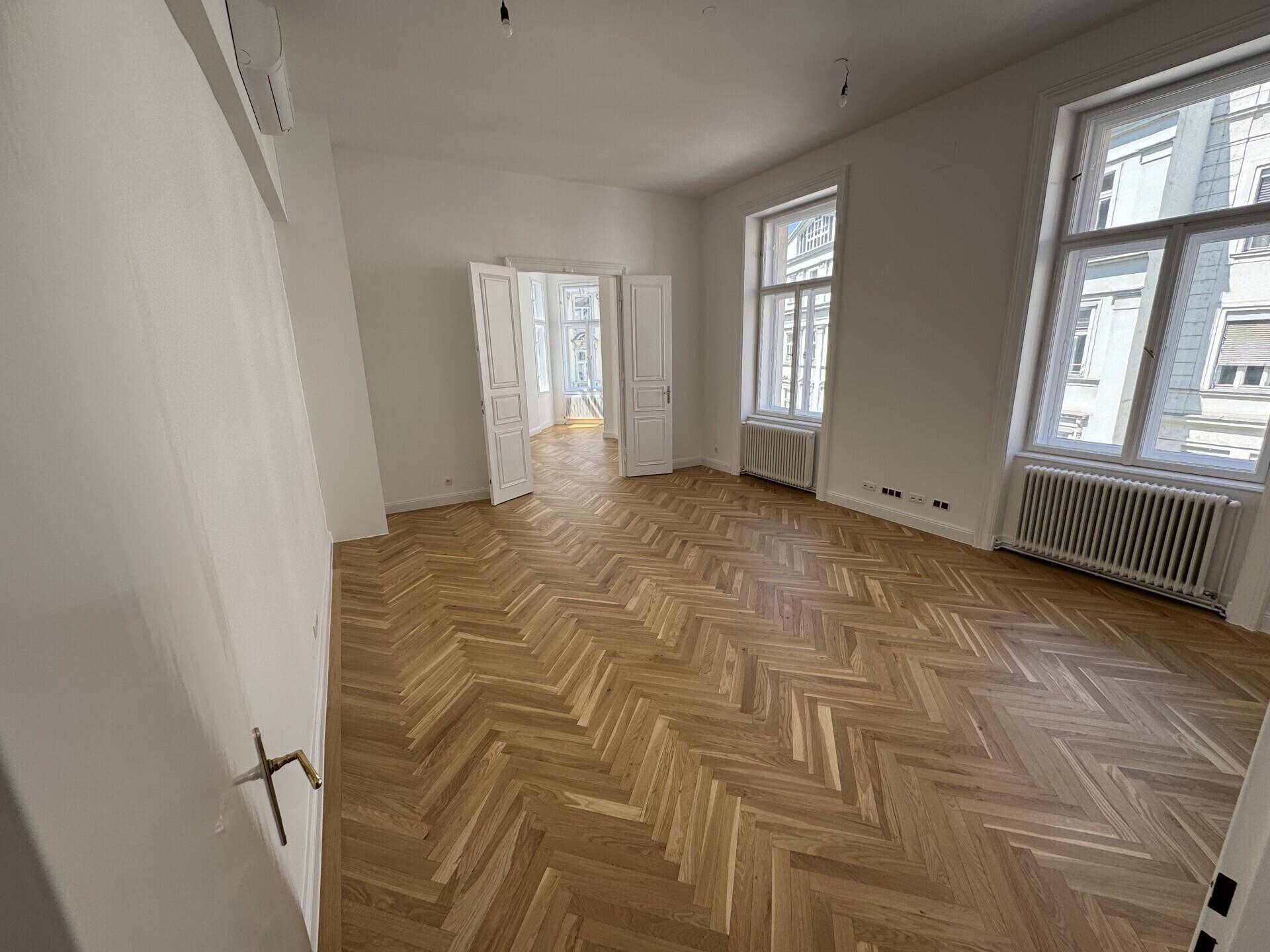 Eleganter Wohnraum mit Fischgrätparkett und Doppeltüren, die zu einem weiteren Zimmer führen.