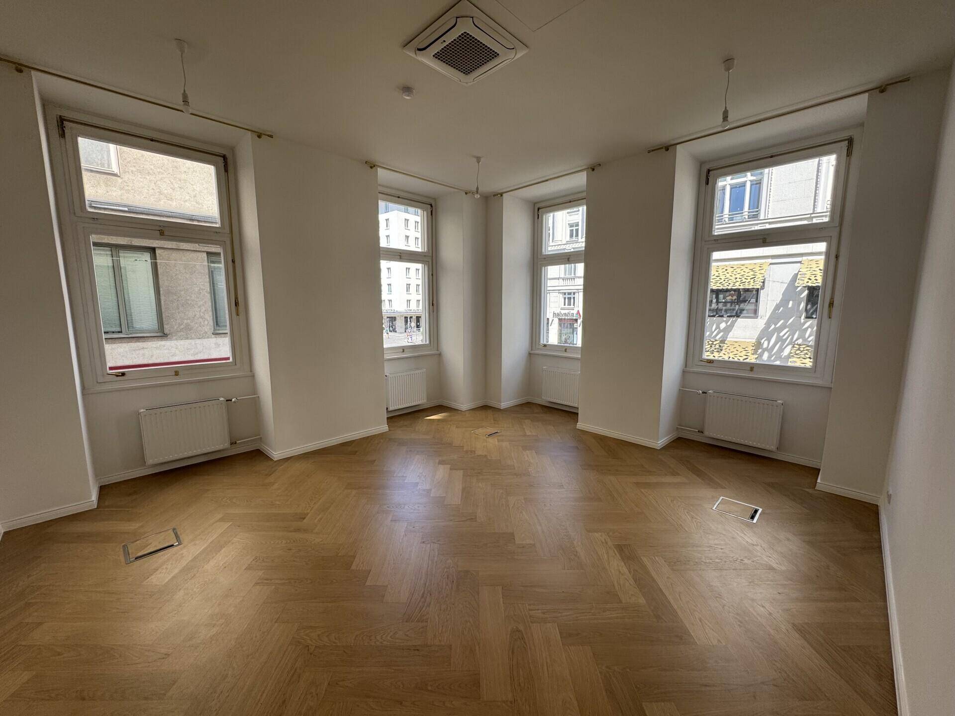 Heller Büroraum mit Fischgrätparkett, hohen Fenstern und Klimaanlage an der Decke.