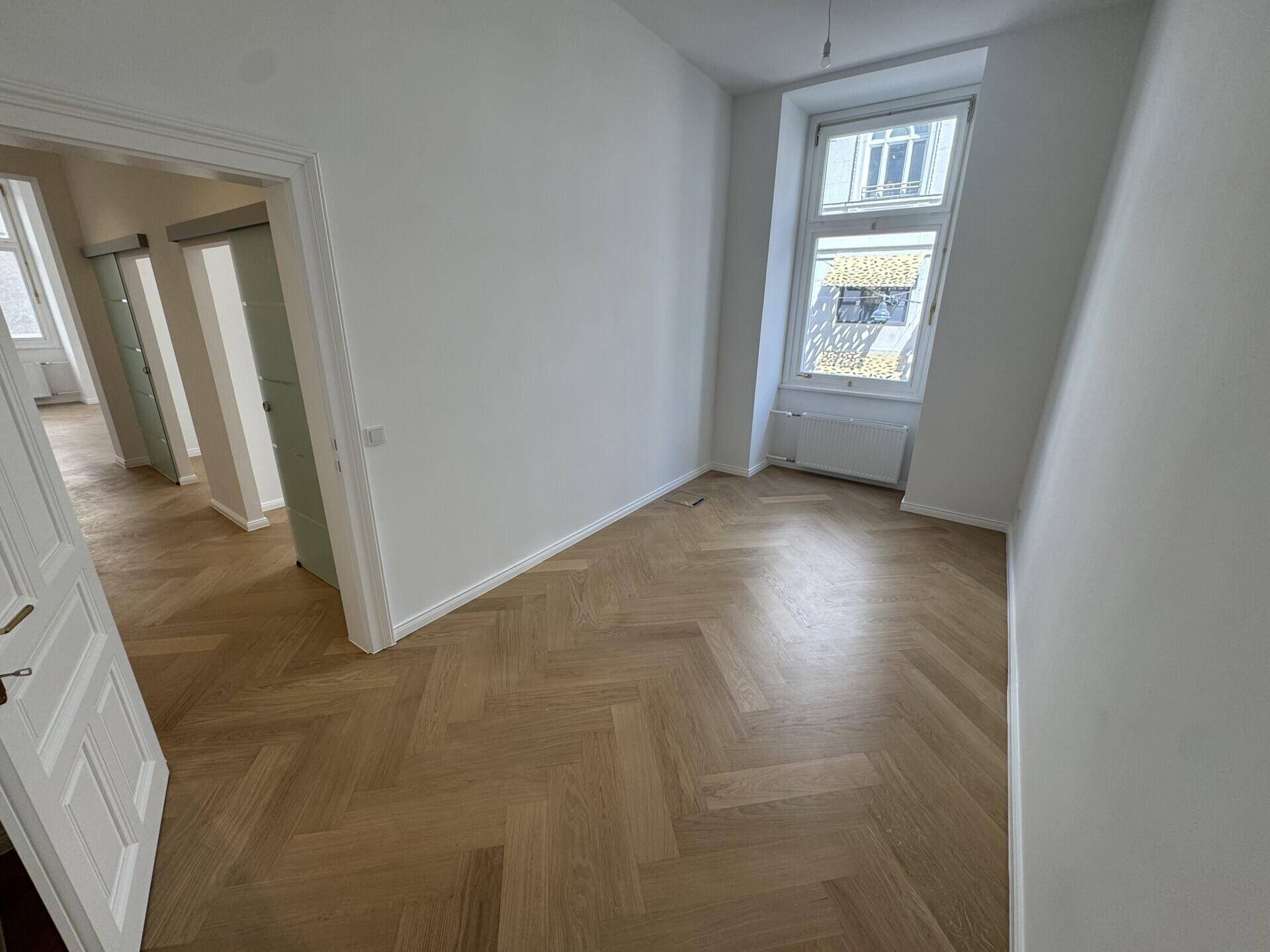 Leerer Büroraum mit hellem Fischgrätparkett, weißen Wänden und einem großen Fenster.