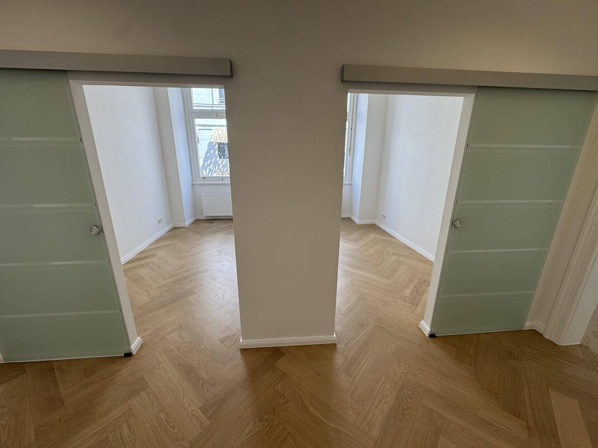 Zwei helle Büroräume, getrennt durch moderne Glasschiebetüren, mit Fischgrätparkett.