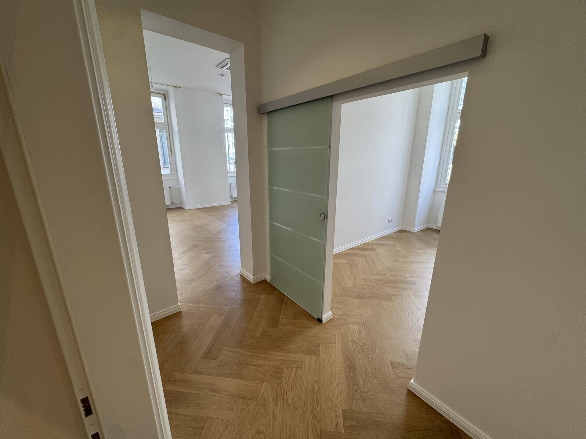 Heller Flur mit Fischgrätparkett, der zu einem Büroraum mit Glasschiebetür führt.