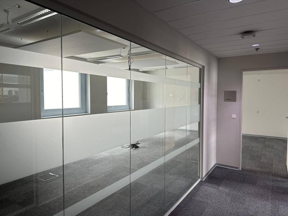 Moderner Bürobereich mit Glasabtrennung und grauem Teppichboden, der viel Licht durchlässt.