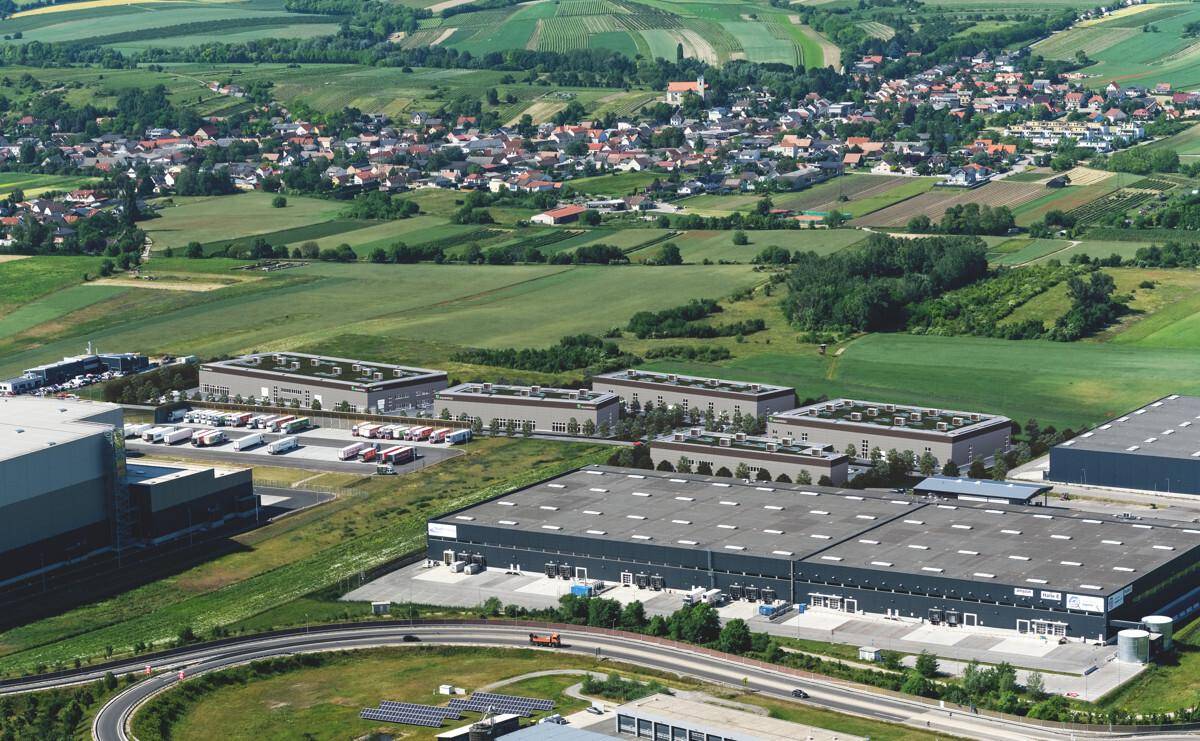 Luftaufnahme eines Industriegebiets mit modernen Hallen, Parkplätzen und angrenzenden Feldern und Wohngebieten.