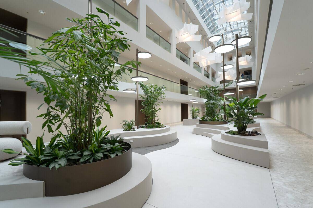 Helles Atrium mit hohen Decken, Glasdach und üppiger Bepflanzung in modernen Pflanzgefäßen.