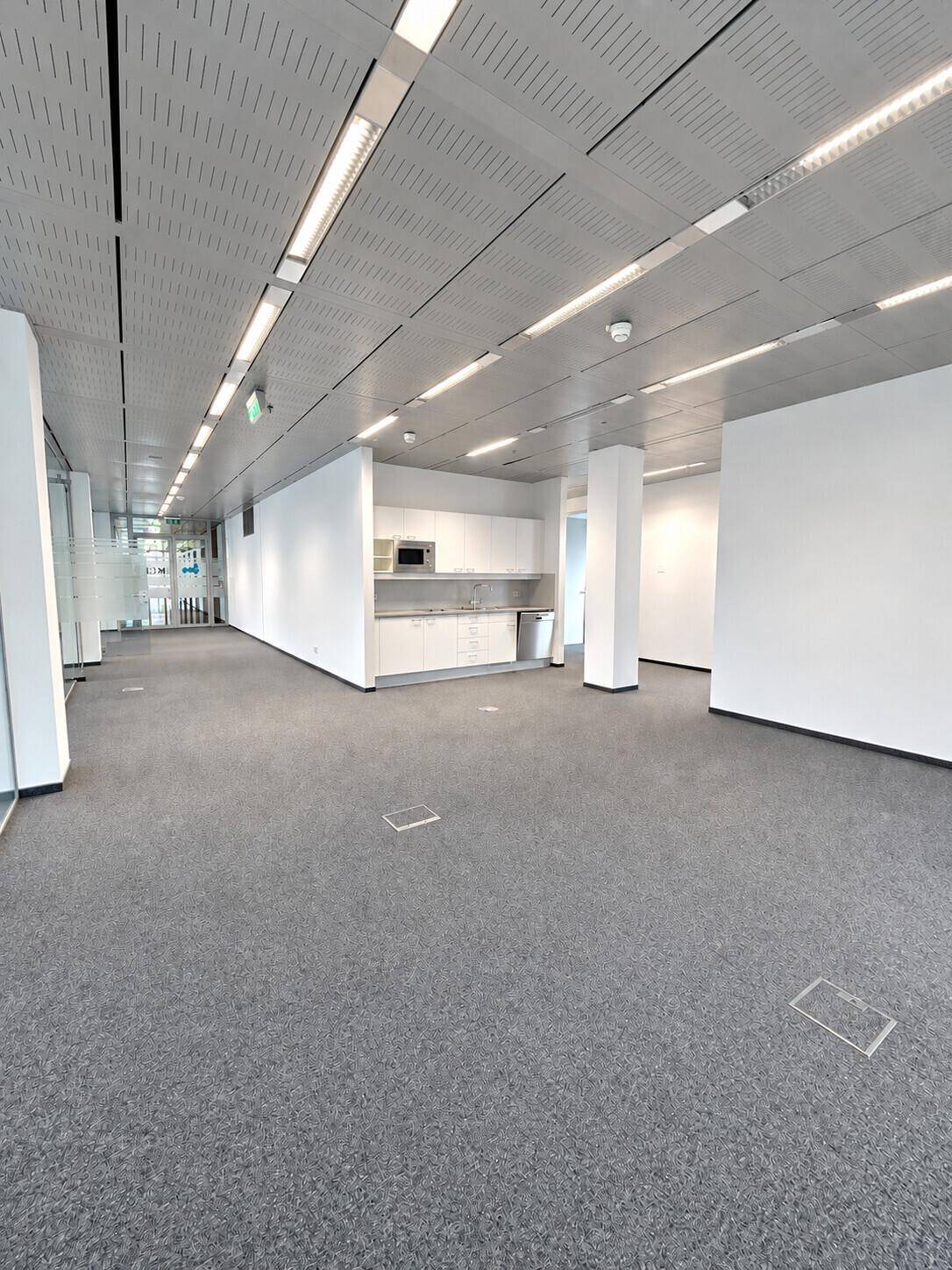 Langer Bürogang mit Teppichboden, weißen Wänden und einer integrierten Küchenzeile.