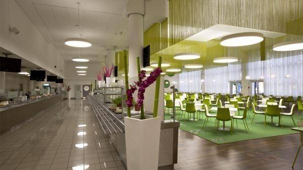 Große Kantine mit vielen Tischen und Stühlen, heller Beleuchtung und grünen Akzenten.
