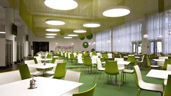 Geräumige Kantine mit vielen Sitzgelegenheiten, grünen Teppichböden und runden Deckenleuchten.