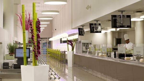Servierbereich einer modernen Kantine mit langen Theken, Bildschirmen und dekorativen Pflanzen.