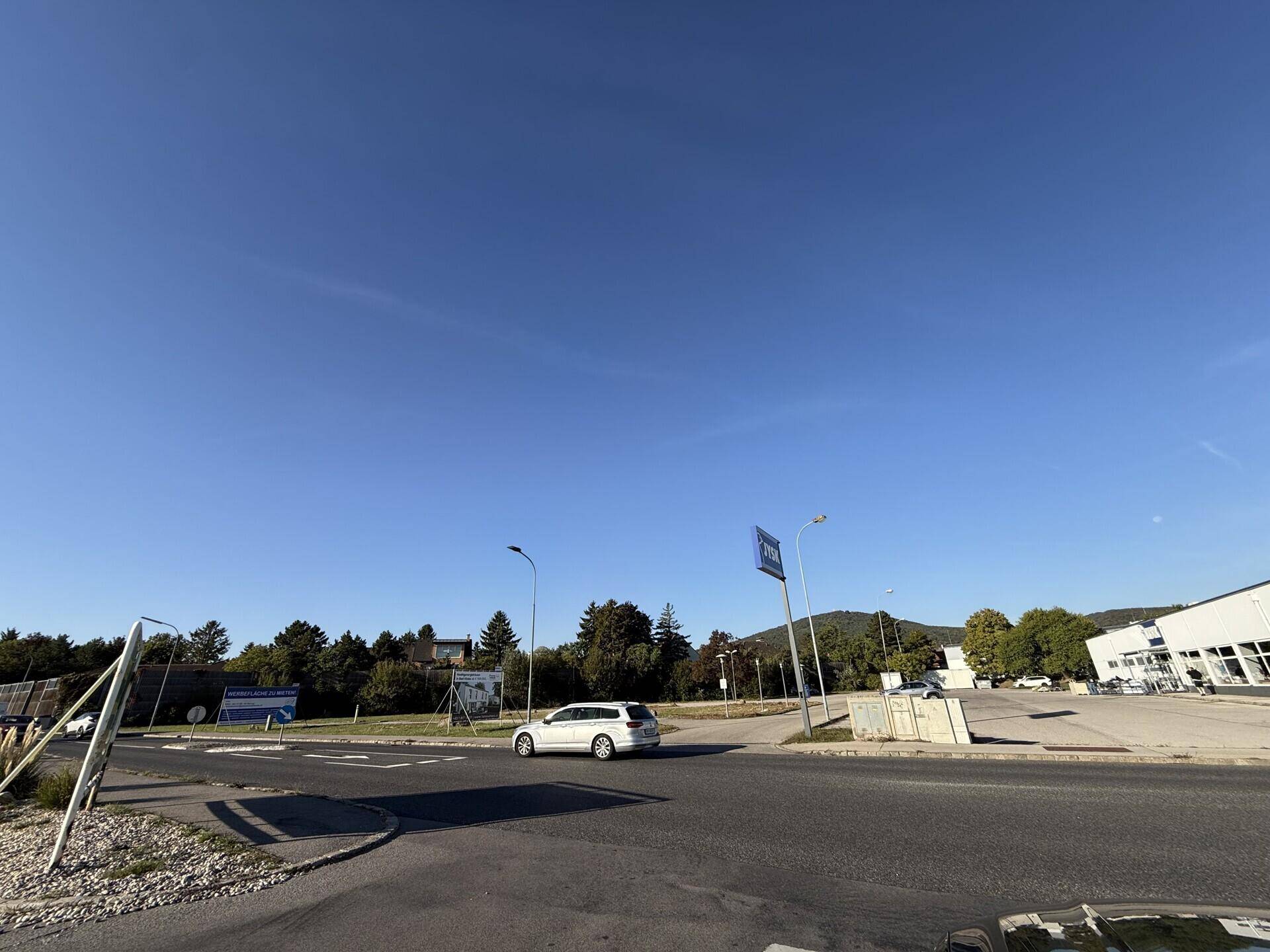 Eine breite Außenaufnahme mit einer Straße, einem fahrenden Auto und einem Parkplatz unter strahlend blauem Himmel.