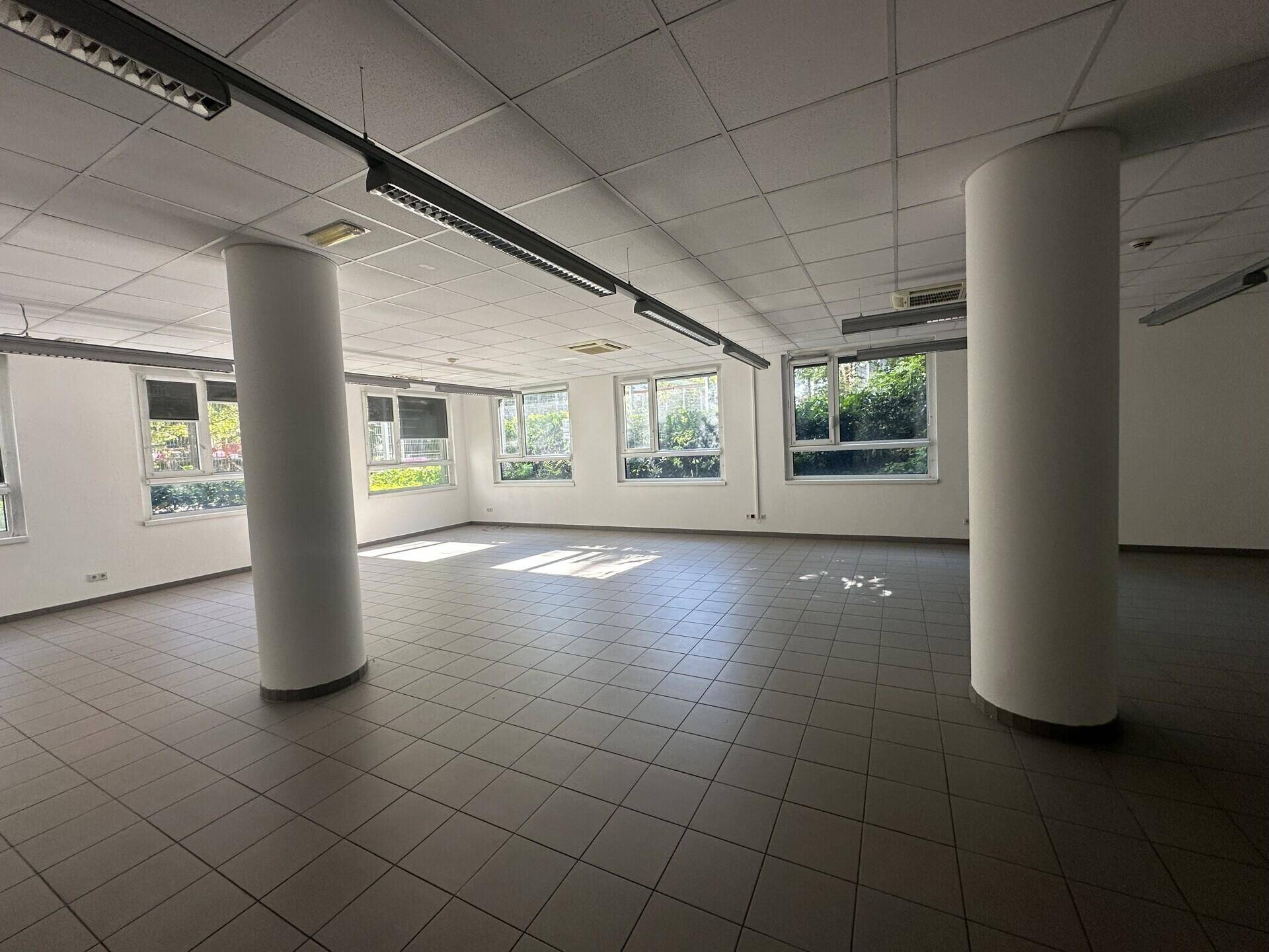 Geräumiger, leerer Büroraum mit Fliesenboden, Säulen und vielen Fenstern, die Tageslicht hereinlassen.