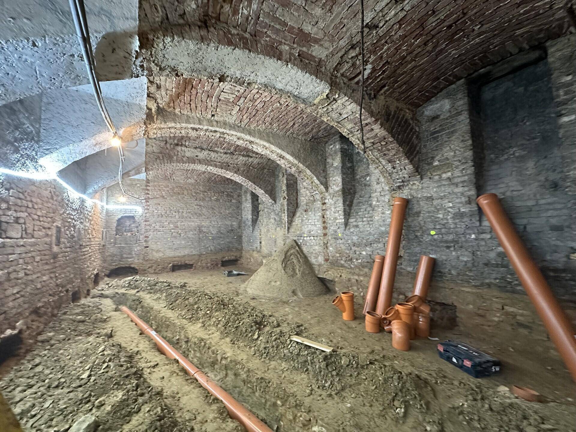 Alter Kellerraum mit Ziegelgewölben und Erdboden, vorbereitet für Bauarbeiten mit Rohren und Sand.