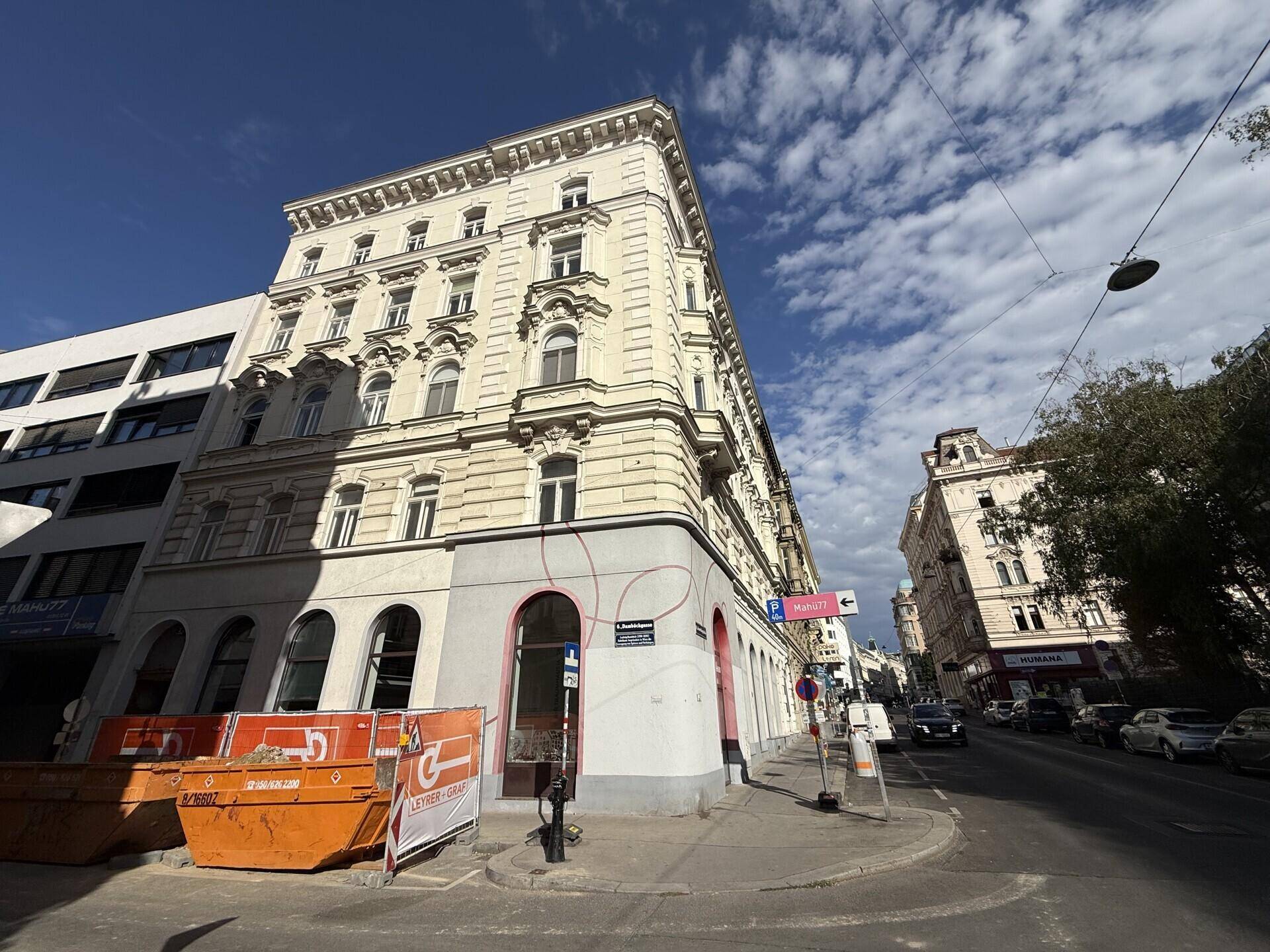 Eckgebäude mit historischer Fassade und Baustellencontainern im Vordergrund unter einem bewölkten Himmel.