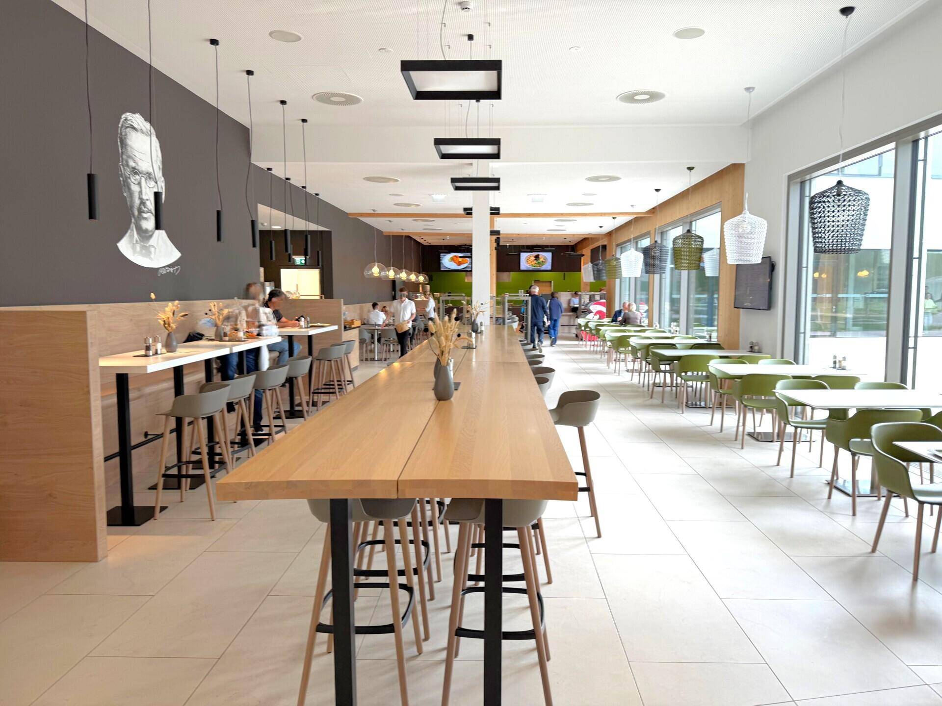 Helle Cafeteria mit verschiedenen Sitzbereichen, langer Theke und moderner Beleuchtung.