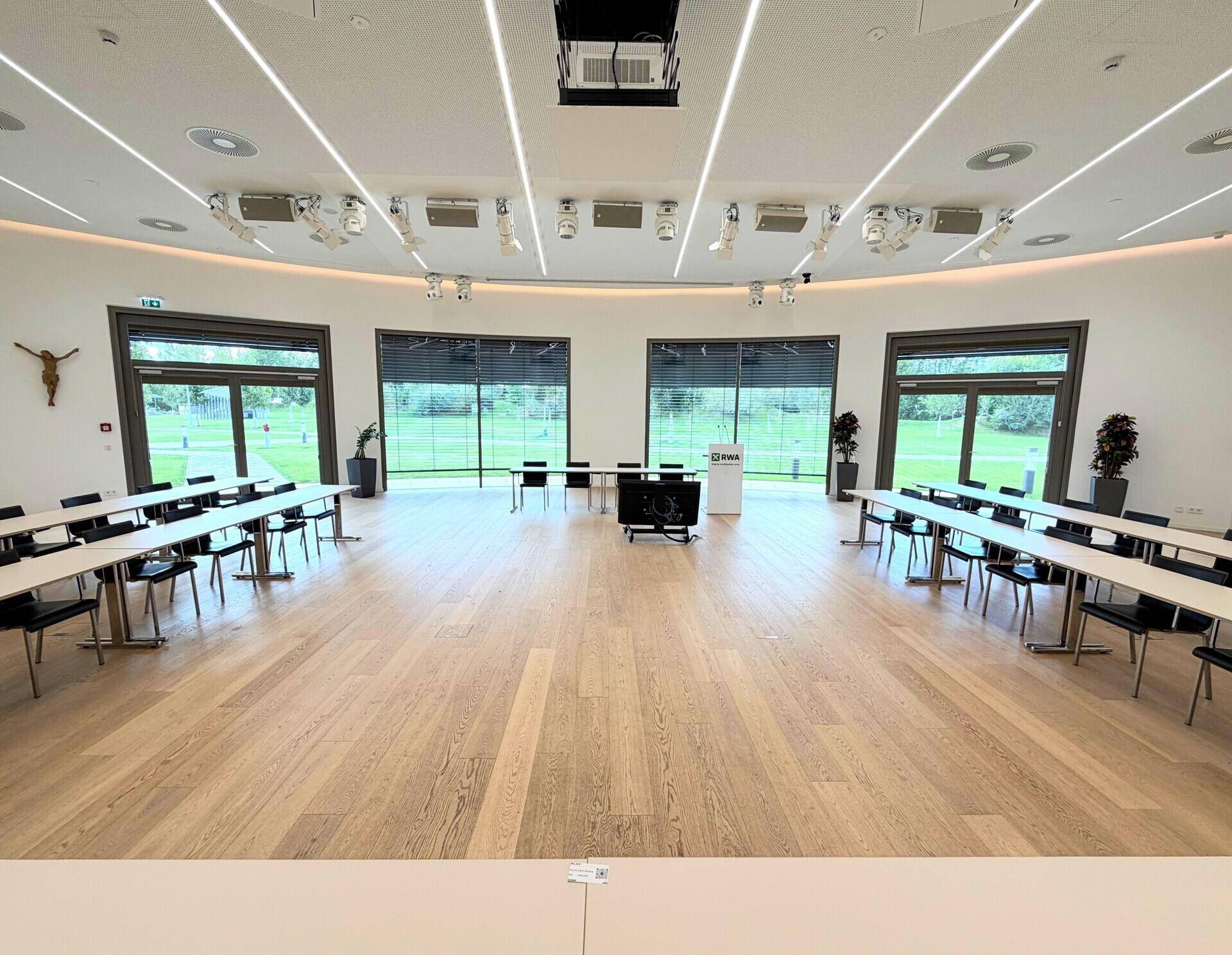 Großer Konferenzsaal mit vielen Tischen und Stühlen, umgeben von großen Fenstern.