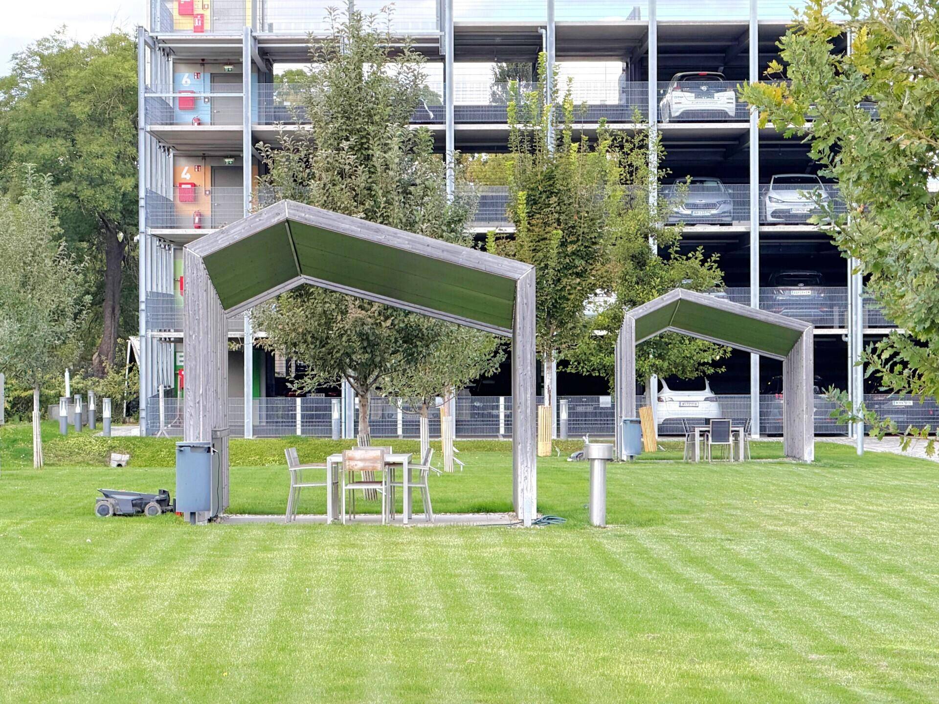 Grüne Außenanlage mit überdachten Sitzbereichen und einem Parkhaus im Hintergrund.