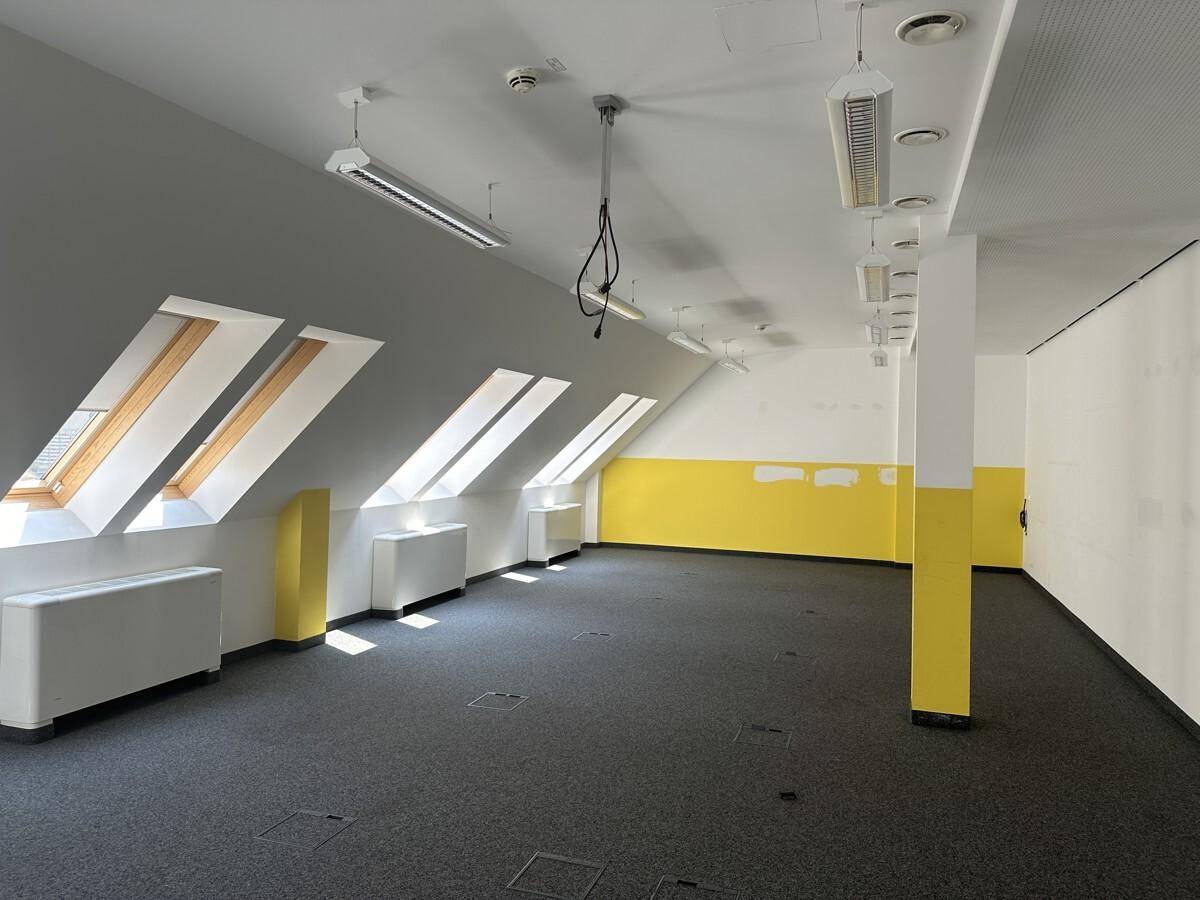 Großer Büroraum im Dachgeschoss mit Teppichboden, gelben Akzentwänden und mehreren Dachfenstern.