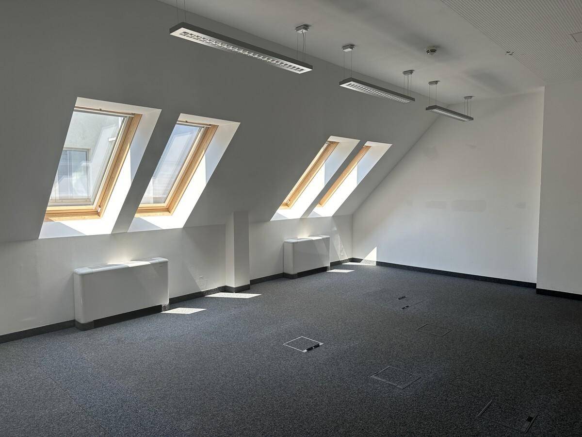 Heller Büroraum im Dachgeschoss mit Teppichboden, schrägen Wänden und mehreren Dachfenstern.