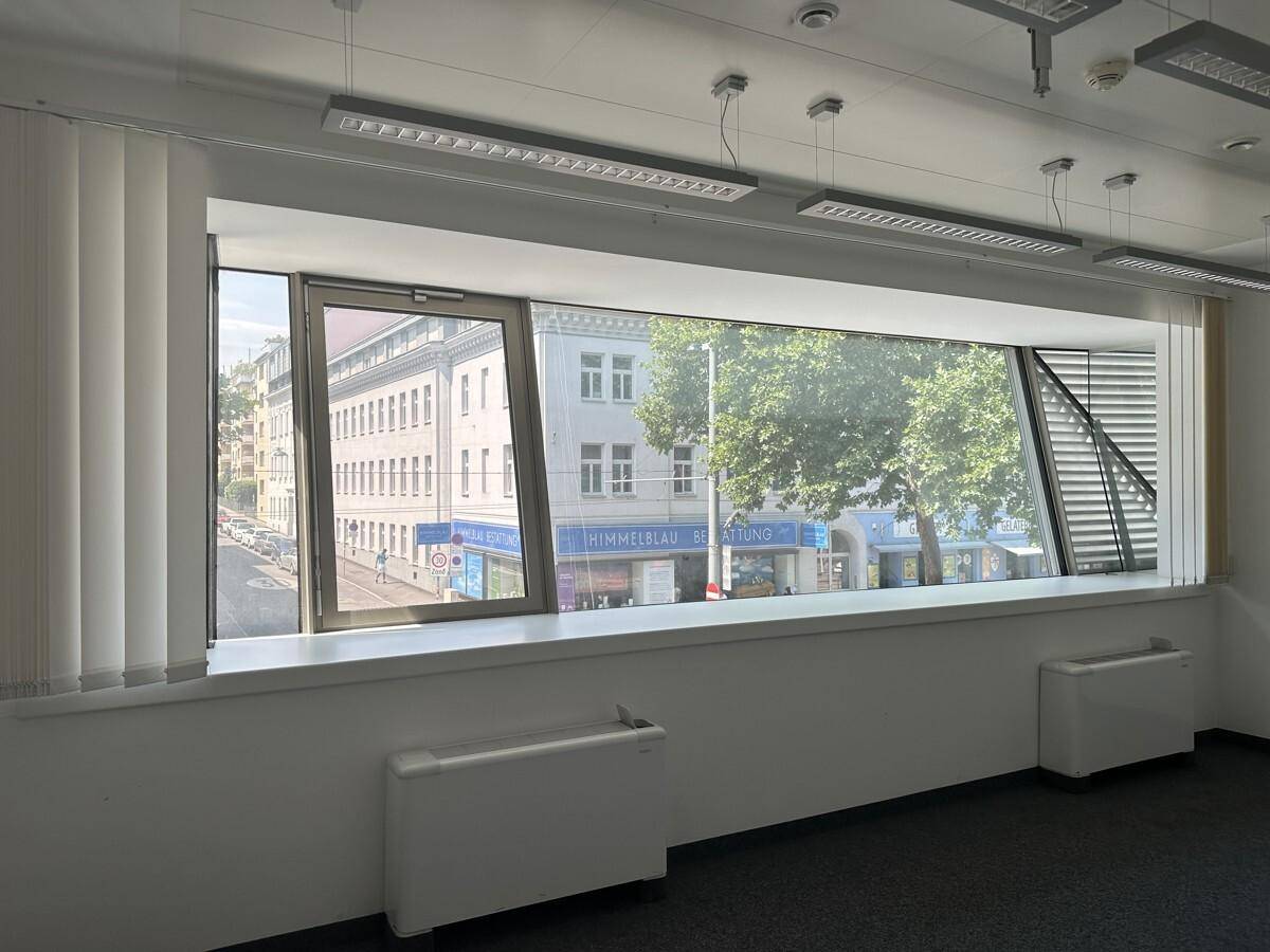 Heller Büroraum mit Teppichboden, breiten Fenstern und Blick auf die belebte Straße und Gebäude.