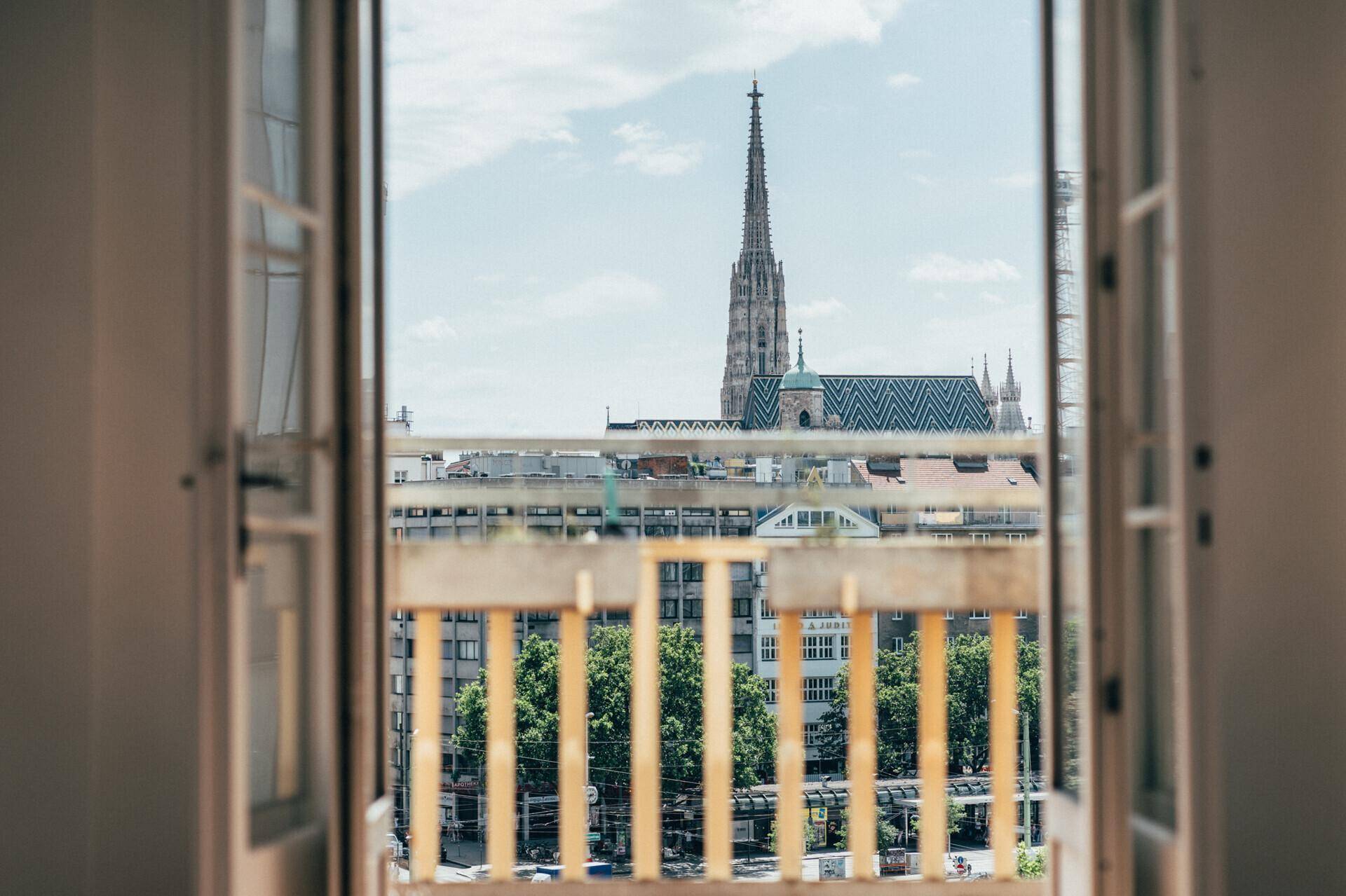 Blick aus einem Fenster oder Balkon auf eine historische Kirche und die Dächer der Stadt unter einem bewölkten Himmel.