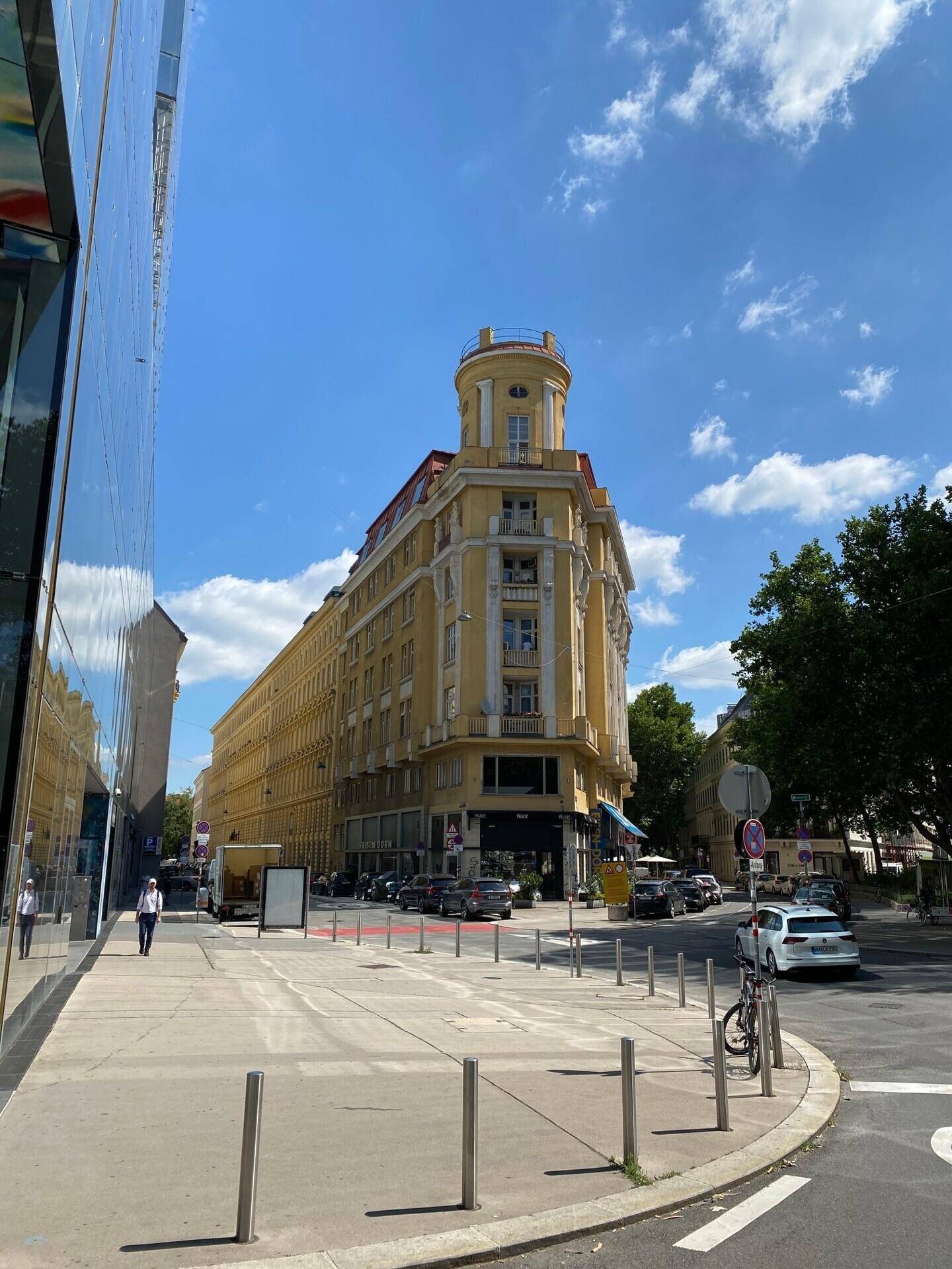 Straßenansicht eines prächtigen gelben Eckgebäudes mit klassischer Architektur und einem runden Turm unter blauem Himmel.