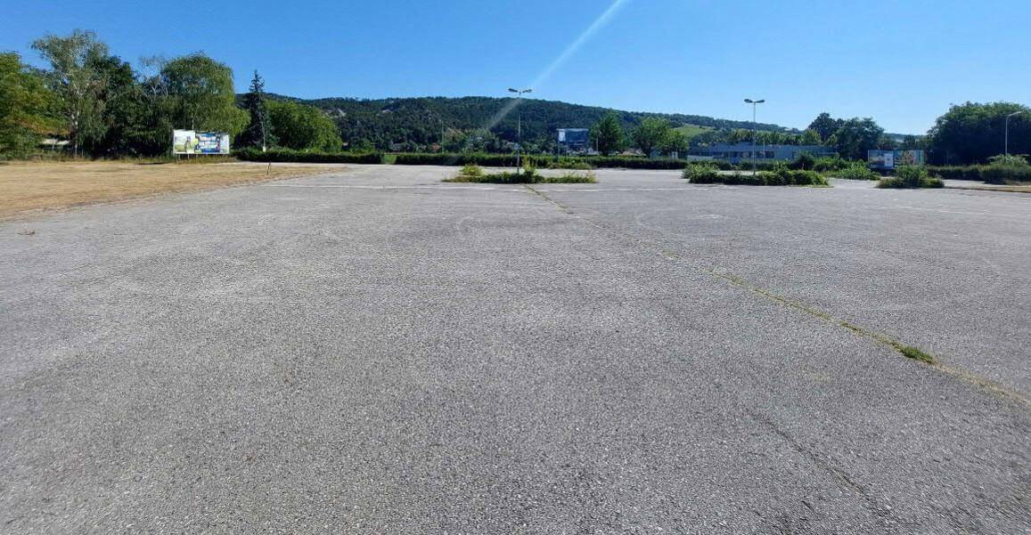 Großer leerer Parkplatz aus Asphalt mit Bäumen und einer Hügellandschaft am Horizont.