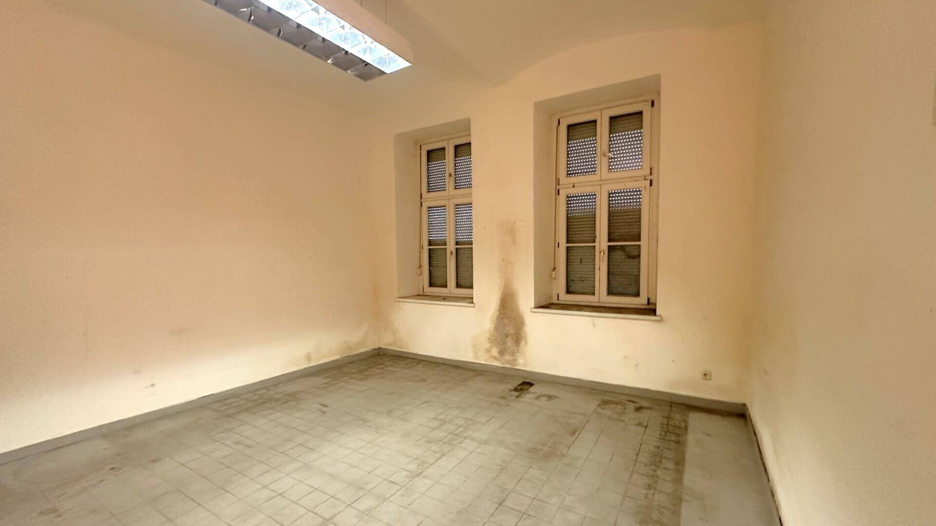 Ein leerer Raum mit hellen Wänden, zwei Fenstern mit Jalousien und sichtbaren Feuchtigkeitsflecken an der Wand.